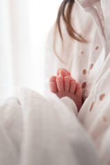 Neonato in braccio alla mamma. si vedono solo i piedini che spuntano da un leggero lenzuolino bianco. i piedini puntano sul petto della mamma, che indossa una camicia in sangallo. della mamma si vede solo la camicia e un ciuffo di capelli