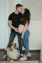 Giovane coppia in dolce attesa. La mamma indossa jeans e top nero, il papà jeans neri e maglietta nera. Fra i piedi del papà c'è il loro cane, pastore australiano. loro si stanno baciando, il cane sbadiglia vistosamente. Il papà sta accarezzando delicatamente la pancia della mamma