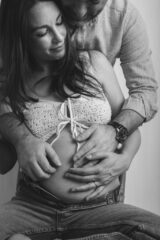 immagine maternity bianco e nero, focus sulla mamma e pancia. mamma seduta con gambe piegate, jeans e piccolo top all'uncinetto. papà che entra nella foto dall'alto accarezzando la pancia della futura mamma. sorrisi a fior di labbra e occhi chiusi, mani che si toccano sulla pancia. foto in bianco e nero