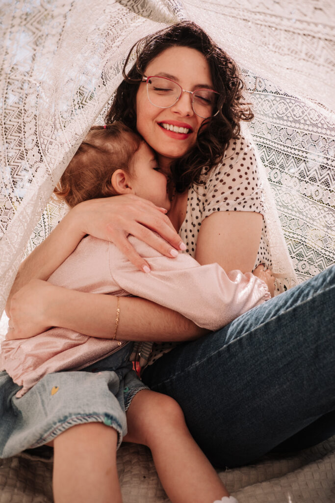 mamma e bimba abbracciate sotto un leggero telo di pizzo che lascia passare la luce del mattino, colori tenuti, rosa e jeans