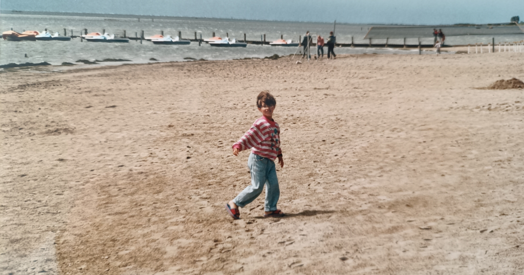 sveva piccola al mare, con felpa a righe bianche e rosa e jeans chiari e ciabatte, in primavera. spiaggia vuota, mare in lontanaza,