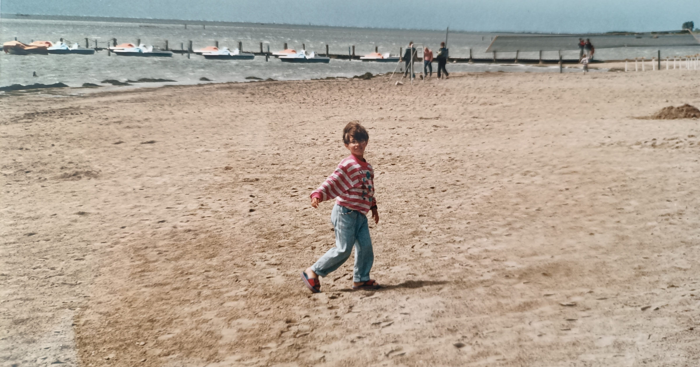 sveva piccola al mare, con felpa a righe bianche e rosa e jeans chiari e ciabatte, in primavera. spiaggia vuota, mare in lontanaza,