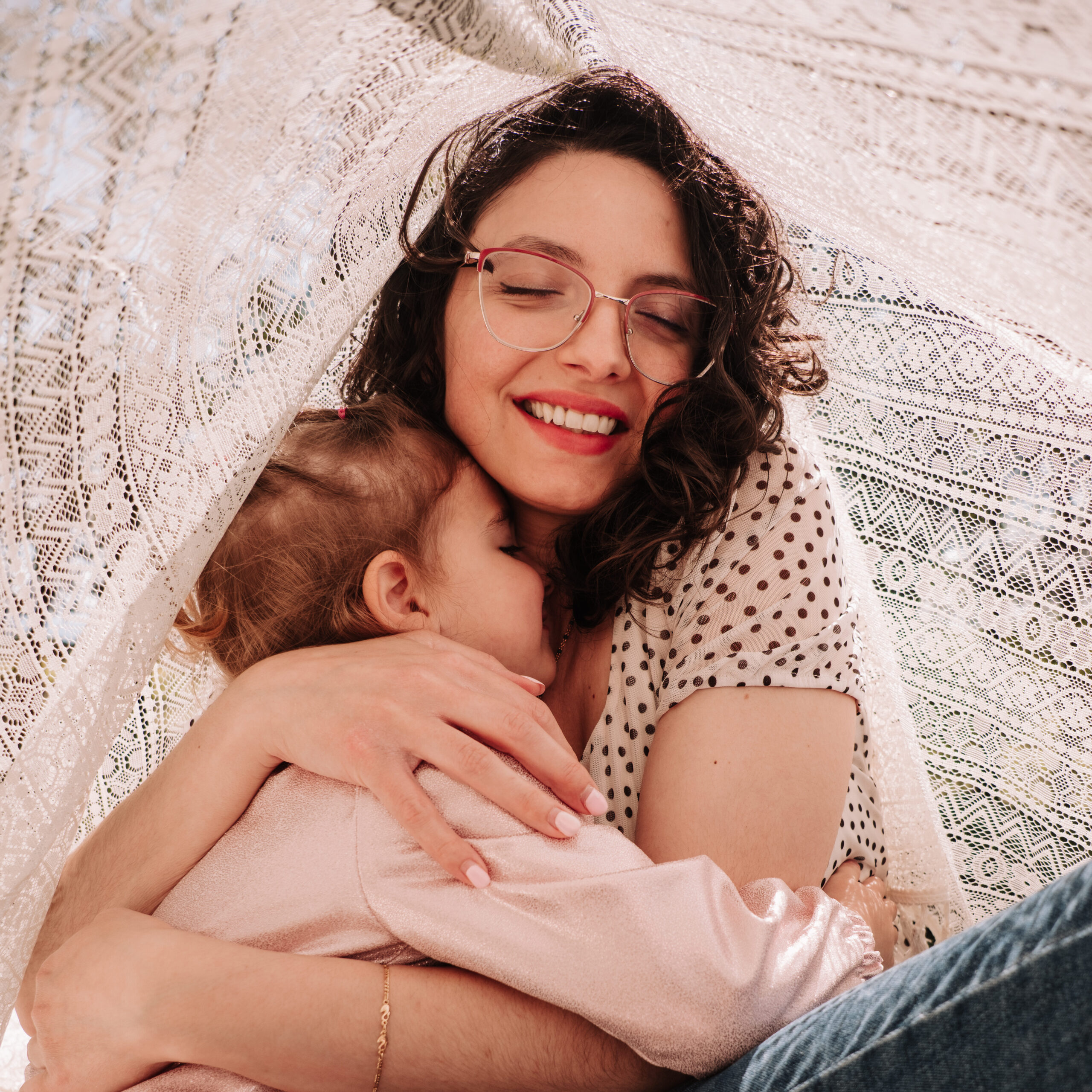 mamma e bimba abbracciate sotto un leggero telo di pizzo che lascia passare la luce del mattino, colori tenuti, rosa e jeans